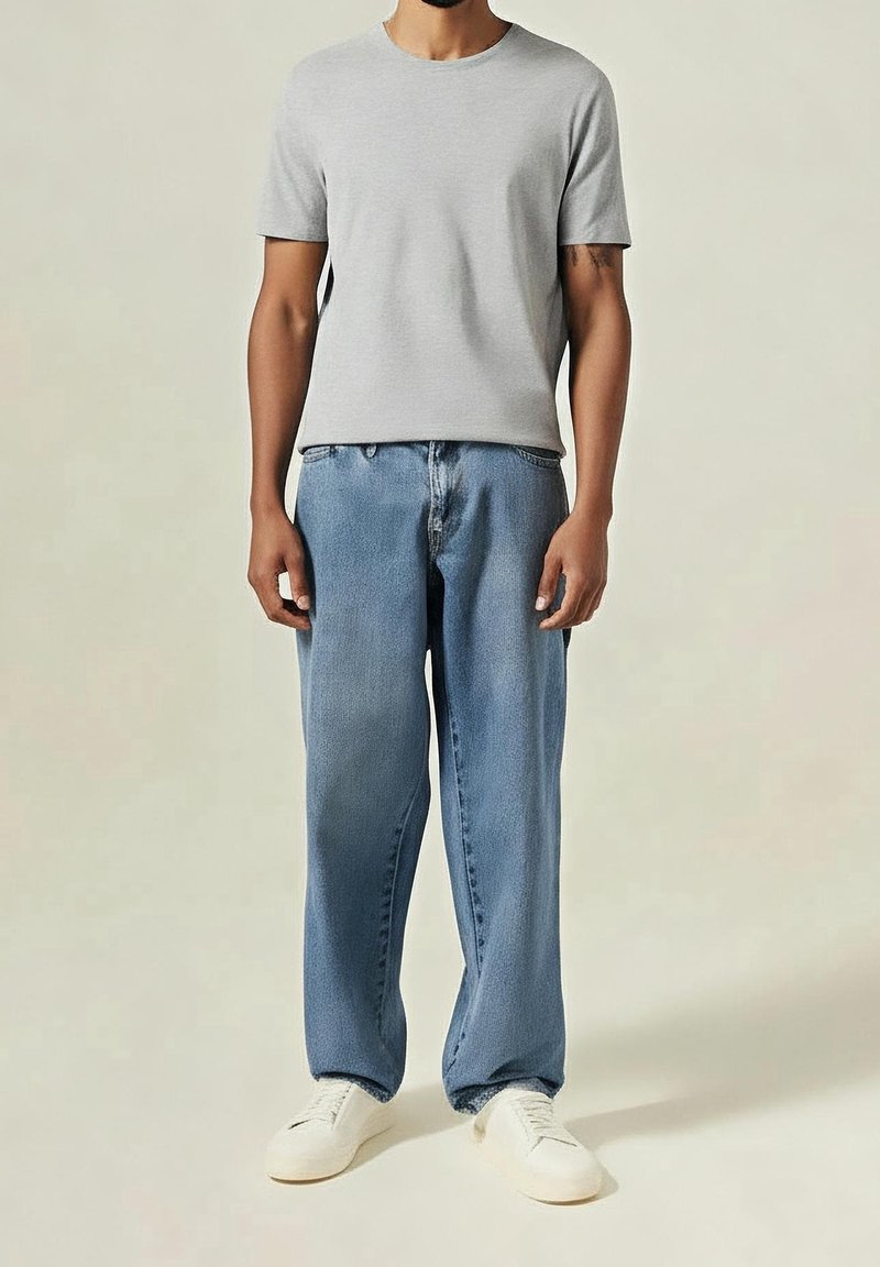 Homme portant un t-shirt gris clair uni, un jean bleu ample et des baskets blanches, debout devant un fond uni clair.