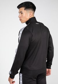 Zwarte zip-up sportjack met hoge kraag, voorzien van witte zijkanten met tekst. Glad textuur, getailleerd ontwerp en geribbelde manchetten.