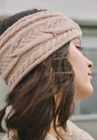 Bentiță tricotată cu model în relief din lână moale bej, cu impletituri texturate și design larg, purtată peste păr întunecat și ondulat.