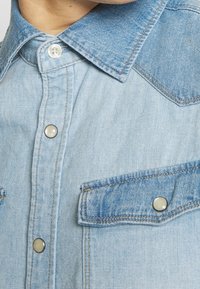 Jasnoniebieska koszula dżinsowa z kołnierzykiem typu button-down, zatrzaskami oraz dwoma kieszeniami na piersi z klapami. Miękka tekstura i klasyczny design.
