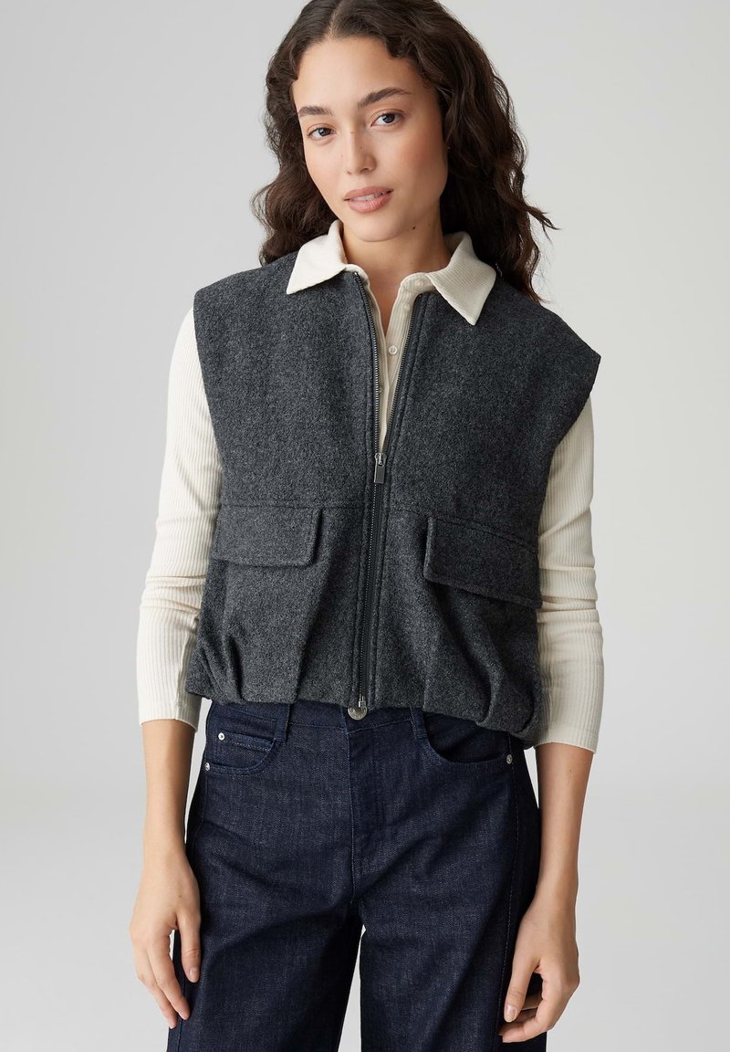 Gilet in lana grigia con colletto alto, cerniera frontale e due tasche applicate. Indossato sopra una maglia a maniche lunghe a costine di colore chiaro. Pantaloni in denim scuro.
