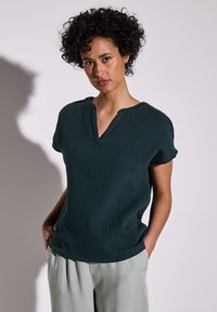 Blusa verde scuro a maniche corte con scollatura a V, tessuto strutturato e vestibilità comoda. Abbinata a pantaloni grigi chiari a gamba larga.