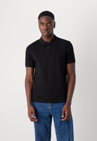 Schwarzes Poloshirt aus einem weichen Baumwollmix mit einer Drei-Knopf-Leiste, kurzen Ärmeln und dezentem besticktem Logo. Kombiniert mit blauen Jeans.