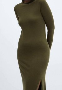 Femme portant une robe midi ajustée à manches longues de couleur vert olive, avec une fente latérale, debout devant un fond clair uni.