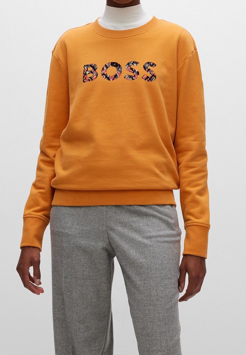 Personne portant un sweat-shirt orange avec le texte "BOSS" à motif floral sur un col roulé blanc et un pantalon gris, debout devant un fond uni.