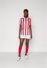 Fodboldtrøje med røde og hvide lodrette striber, med en logo på brystet. Kombineret med hvide shorts og røde sokker. Sportsko er synlige.