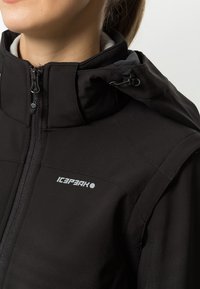 Giacca softshell nera con zip frontale, collo alto e cappuccio regolabile. Presenta un logo sul petto e tessuto strutturato.