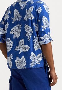 Niebieska koszula z krótkim rękawem z białym tropikalnym wzorem liści, wykonana z lekkiego materiału, z podwiniętymi rękawami i luźnym krojem.