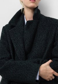 Cappotto in lana verde scuro, texturizzato, con colletto a risvolto e design oversize. Presenta un ampio revers e copre la parte superiore del corpo con maniche lunghe.