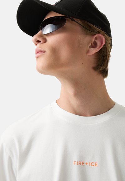 White cotton T-shirt with round neckline and orange "FIRE+ICE" text. Black cap and dark sunglasses complete the look.