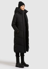 Abrigo de plumas negro con cuello alto, cierre de cremallera, bolsillos laterales y detalles a presión. Combinado con botas negras hasta la rodilla y un gorro.