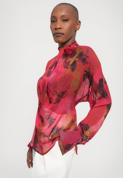 Doorzichtig, rode blouse met bloemmotieven in tinten van roze, oranje en zwart. Kenmerken zijn een hoge kraag en lange mouwen.