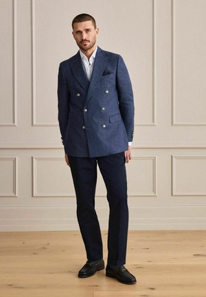 Man wearing navy double-breasted blazer, white shirt, dark trousers, and black loafers standing in a beige room with wooden floor.