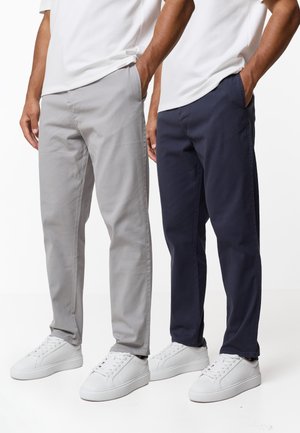 Deux hommes portant des baskets blanches et des pantalons décontractés, l'un en gris clair et l'autre en bleu marine foncé, tous deux les mains dans les poches.
