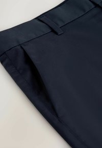 Gros plan sur un pantalon bleu marine montrant la ceinture avec passants et l'ouverture de la poche latérale sur un fond clair.