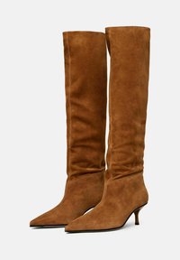 Botas de rodilla de ante marrón con punta afilada y un tacón bajo y estrechado. El diseño presenta una silueta ajustada y una costura mínima.