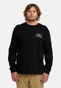 Maglietta nera a maniche lunghe in cotone con una piccola toppa logo colorata sul petto. Presenta polsini a costine e un collo a girocollo.