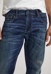 Jeans de mezclilla en un color índigo profundo con detalles desgastados, diseño de cinco bolsillos y costuras en contraste a lo largo de las costuras y la cinturilla.