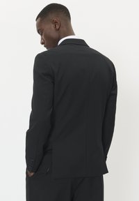 Melna pielāgota uzvalka žakete ar vienu ventilāciju, mīkstajiem uzgaļiem un pogātām piedurknēm. Gluda auduma, strukturēts dizains.