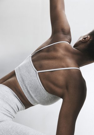 Vrouw in grijze sportkleding stretcht met opgeheven arm, toont rugspieren tegen een effen lichte achtergrond.