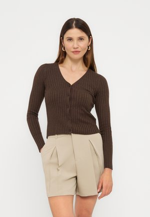 Donna con lunghi capelli castani che indossa orecchini a cerchio dorati, un cardigan marrone a coste con bottoni e pantaloncini beige plissettati, in piedi con le mani nelle tasche.