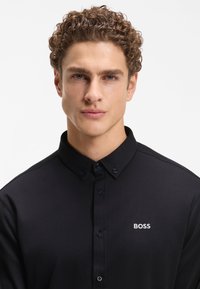 Μαύρο πουκάμισο με γιακά από υφασμάτινη υφή, με μικρό λευκό λογότυπο "BOSS" στην αριστερή πλευρά και σκούρα κουμπιά στο μπροστινό μέρος.
