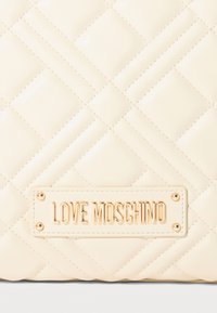 Κρεμ καπιτονέ δερμάτινη επιφάνεια με χρυσή μεταλλική πλάκα που γράφει "LOVE MOSCHINO", προσαρτημένη με τέσσερις χρυσούς κοχλίες.