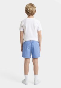 Παιδί που στέκεται με την πλάτη στραμμένη, φοράει λευκό μπλουζάκι, ανοιχτό μπλε σορτς, λευκές κάλτσες και λευκά αθλητικά παπούτσια σε απλό λευκό φόντο.