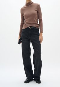 Personne portant un col roulé marron, un jean noir à jambes larges avec ceinture, des mocassins noirs, tenant une pochette noire sur fond blanc.