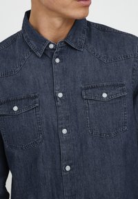 Camisa de denim oscuro con cuello puntiagudo, dos bolsillos en el pecho y botones blancos. La tela tiene una textura suave y un corte clásico.
