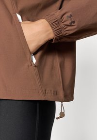 Καφέ μπουφάν με ελαστικές μανσέτες, πλαϊνές τσέπες και ρυθμιζόμενο κάτω μέρος με κορδόνι. Διαθέτει λογότυπο στο μανίκι και έχει μαλακή, ματ υφή.