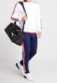 Biała bluza z czerwonymi i czarnymi paskami, granatowe dresy z czerwonymi i białymi paskami po bokach oraz czarna torba sportowa. Białe buty sportowe.
