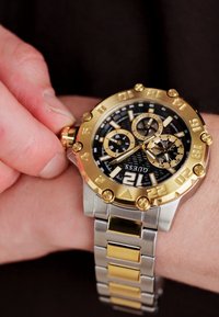 Gold- und Silbermetall-Uhr mit schwarzem Ziffernblatt, goldenen Akzenten und großen Ziffern. Verfügt über Chronographen-Subdials und eine strukturierte Lünette.