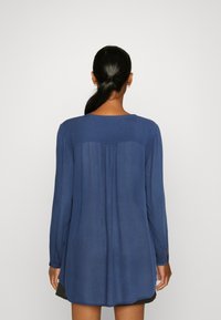 Camicia a maniche lunghe di colore blu navy con scollatura tonda, pieghe sul retro e orlo leggermente più lungo, realizzata in un tessuto fluido.