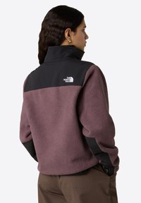 Giacca in pile di un viola tenue con sovrapposizione nera sulle spalle. Presenta un colletto alto e un dettaglio del logo sulla parte superiore della schiena. Texture morbida.