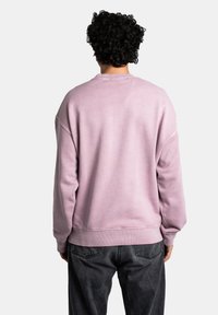 Felpa rosa pastello con colletto a girocollo, spalle scese e polsini a costine. Indossata con jeans grigio scuro, che mostrano una vestibilità rilassata e una texture morbida.
