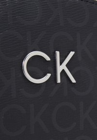 Logo di Calvin Klein su materiale sintetico nero texturizzato. Presenta un prominente insignia argentata "CK" in mezzo a un motivo ripetuto di lettere.