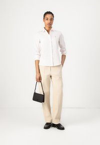 Lichtroze blouse met lange mouwen en knoopsluiting, beige wijde broek, en een kleine zwarte handtas, gecombineerd met zwarte schoenen.