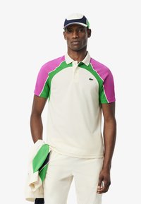 Kortärmad polo i krämfärg med färgglada lila och gröna mesh-paneler, klassisk krage och Lacoste-logga. Tillsammans med en hatt.
