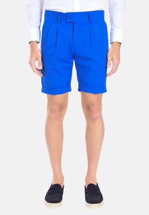 Pantaloni corti blu reale con pieghe, orlo risvoltato e chiusura a bottone. Indossati con una camicia bianca e espadrillas scure.