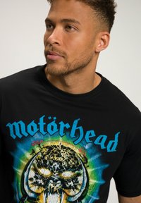 Sort t-shirt med farverigt grafisk print af et krakket kranie; "Motörhead" tekst vist i blåt over designet. Blødt bomuldsstof.