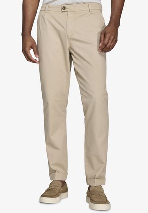 Mężczyzna w beżowych chinosach z podwiniętymi nogawkami i brązowych zamszowych mokasynach stoi na białym tle.