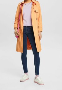 Jasnopomarańczowy płaszcz trench z guzikami, noszony na lawendowej bluzce i niebieskich skinny jeansach. Białe trampki z różowymi akcentami dopełniają stylizację.
