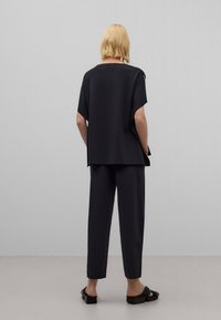 Henkilö, jolla on lyhyet vaaleat hiukset, seisoo selkä kameralle, päällään löysä musta paita, mustat housut ja mustat matalakorkoiset sandaalit harmaata taustaa vasten.