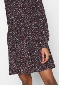 Musta mekko, jossa on vaaleanpunaisia, valkoisia ja ruskeita kukkakuoseja. Varustettu pitkällä hihalla ja rentolla istuvuudella, vyötäröllä on kerroksellista yksityiskohtaa.