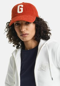 Oranje corduroy pet met een witte geborduurde letter "G" op de voorkant. Draagt een witte ritsvest over een marineblauw shirt.