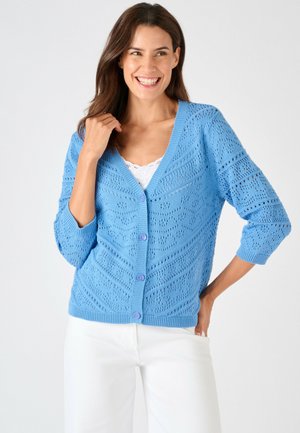 Mujer sonriendo, vistiendo una rebeca de punto azul claro con estampado y botones sobre una camiseta blanca y pantalones blancos sobre un fondo liso.