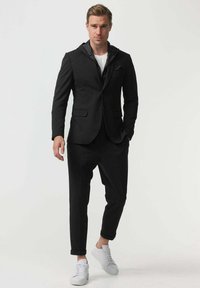 Costum negru cu un sacou structurat care include un hanorac și pantaloni conici, asortați cu adidași albi pe un fundal neutru.