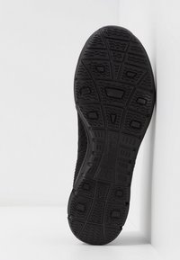 Semelle de basket noire avec des motifs de bande de roulement, comprenant des carrés et des lignes ondulées, positionnée à la verticale contre un mur d’angle blanc.