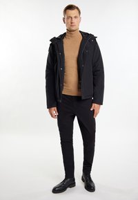 Sort polstret jakke med hætte, båret over en tan turtleneck. Modellen har sorte slim-fit bukser og sorte snørebåndsstøvler på. Rent design.